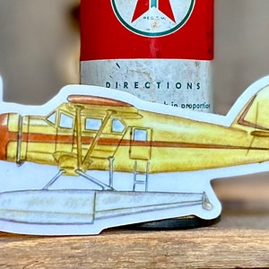 May include: A yellow and brown vintage seaplane sticker with a white background. The seaplane has two floats and a propeller. The sticker is on a red and white can.