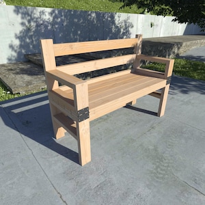 Puede incluir: Un banco de madera con respaldo de listones y apoyabrazos. El banco está hecho de madera natural y tiene un aspecto rústico.