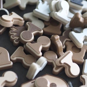 Op de afbeelding: Een verzameling kleine, platte, tweedimensionale spelstukken in diverse vormen, waaronder gestileerde schaakachtige figuren, kronen en geometrische vormen. De stukken zijn uitgevoerd in een palet van matte kleuren: wit, beige, bruin en zwart.