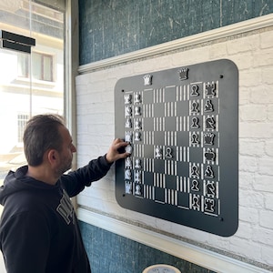 May include: A wall-mounted black chess board with a grid of vertical white lines and rounded corners. The board features magnetic black and white chess pieces arranged in a game. The board is mounted on a white brick-patterned wall surface.