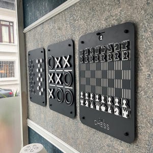 May include: Three black wall-mounted game boards: a dots and boxes game, a tic-tac-toe game, and a chess set. The chess set has white and black pieces. The tic-tac-toe board has white X's and O's. The games are mounted on a patterned wall.