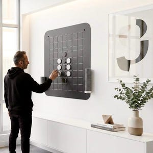 Könnte beinhalten: Ein großes, quadratisches, dunkelgraues, an der Wand montiertes Spielbrett mit einem Gittermuster und runden Spielfiguren in Weiß und Dunkelgrau. An den Seiten des Bretts befinden sich zwei zylindrische Halterungen mit Stapeln weiterer runder Spielfiguren.