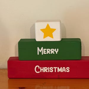 May include: A stack of three rectangular wooden blocks. The bottom block is red with the white text "CHRISTMAS". The middle block is green with the white text "MERRY". The top block is white and features a yellow star graphic.