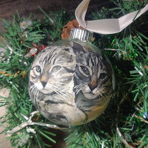May include: A glass Christmas ornament with a hand-painted image of two cats. The cats are a tabby and a calico, and they are looking at the viewer. The ornament is decorated with a white ribbon.