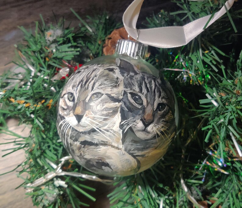 May include: A glass Christmas ornament with a hand-painted image of two cats. The cats are a tabby and a calico, and they are looking at the viewer. The ornament is decorated with a white ribbon.