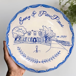 May include: A white ceramic plate with a blue border and a hand-painted design. The design features a house, trees, and the text "Enny & Paw Paws est. 1989".
