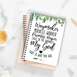 May include: A white notebook with a black spiral binding. The cover features a green leafy design and the text "Waymaker Miracle Worker Promise Keeper Light in the Darkness My God That is who you are".