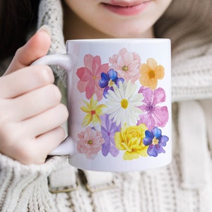 May include: White ceramic mug with a floral design featuring pink, yellow, white, purple, and blue flowers. The flowers are painted in a watercolor style.