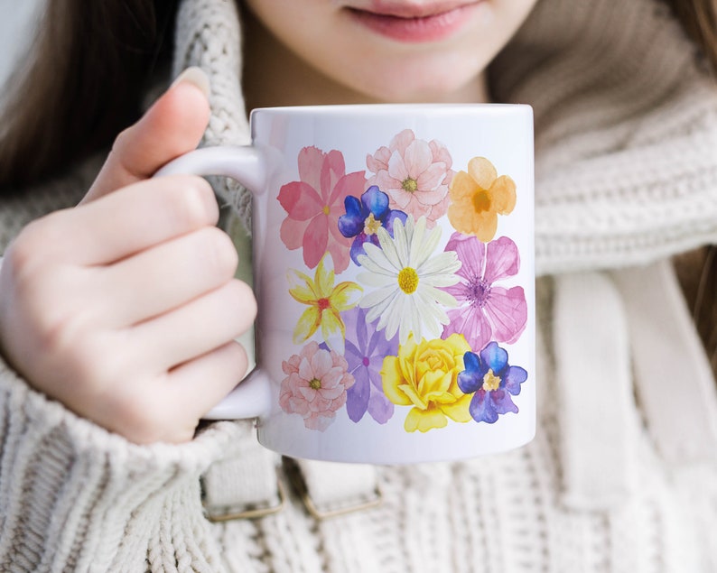May include: White ceramic mug with a floral design featuring pink, yellow, white, purple, and blue flowers. The flowers are painted in a watercolor style.