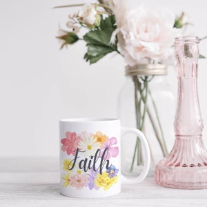 May include: White ceramic mug with a floral design and the word "Faith" in black script. The mug is sitting on a white wooden surface.