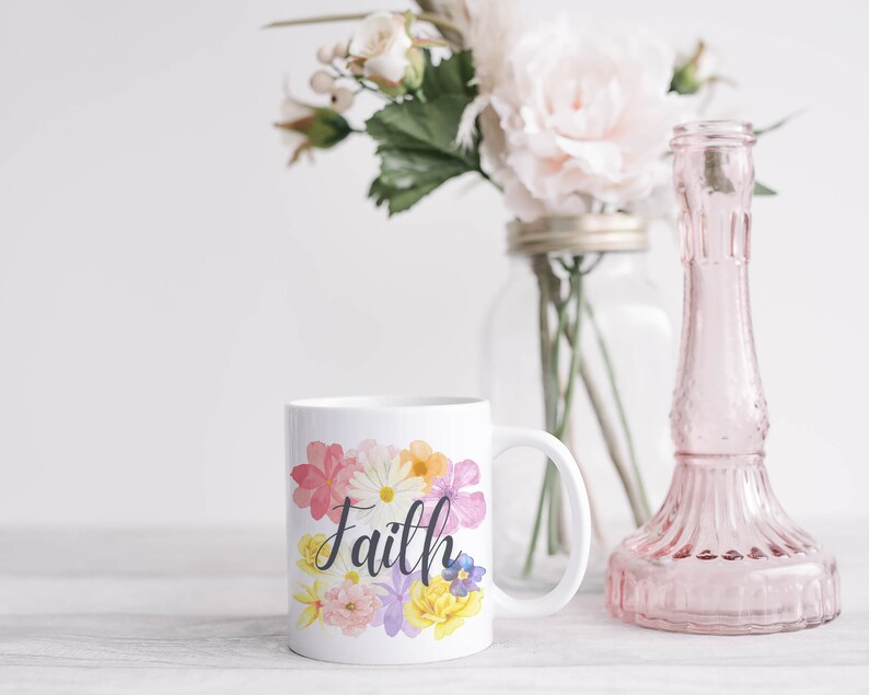 May include: White ceramic mug with a floral design and the word "Faith" in black script. The mug is sitting on a white wooden surface.