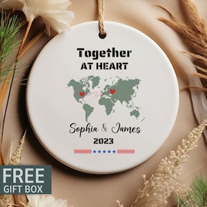 May include: White ceramic ornament with a world map and red hearts connected by a red line. The text "Together at Heart" is at the top, followed by "Sophia & James 2023" and two red, white, and blue stripes with five stars.
