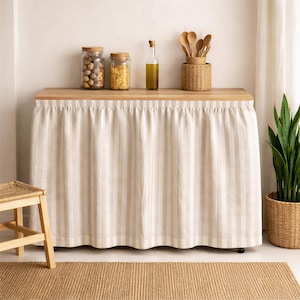 May include: A kitchen island with a light wood countertop and a cream-colored, vertically striped skirt. The countertop holds glass jars with food, a bottle of olive oil, and a woven basket with wooden spoons. A small woven stool and a jute rug are in the foreground.