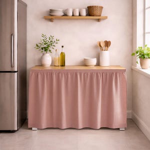 May include: A kitchen island with a light wood countertop and a dusty rose fabric skirt. The island is in a kitchen with a stainless steel refrigerator, a wooden shelf with dishes, and a window with a potted plant.