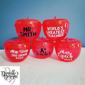Puede incluir: Cinco recipientes rojos en forma de manzana con texto negro. Los recipientes dicen "Mr. Smith", "World's Greatest Teacher", "Mrs. Weiss 3rd Grade 2022", "A+ Teacher", y "Miss Lynch 2022".