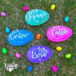 May include: Four colorful plastic Easter eggs with bunny ears and names "Ryan", "Carter", "Callie", and "Kara" written on them. The eggs are on a green grassy surface with several small colorful plastic eggs scattered around.