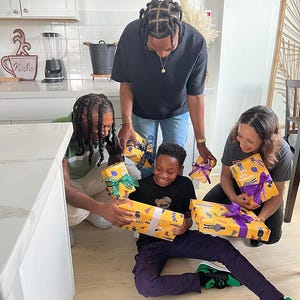 May include: A group of people are gathered, holding and admiring wrapped gifts. The gifts are wrapped in yellow paper with a repeating pattern and tied with colorful ribbons. The scene takes place in a well-lit kitchen setting.