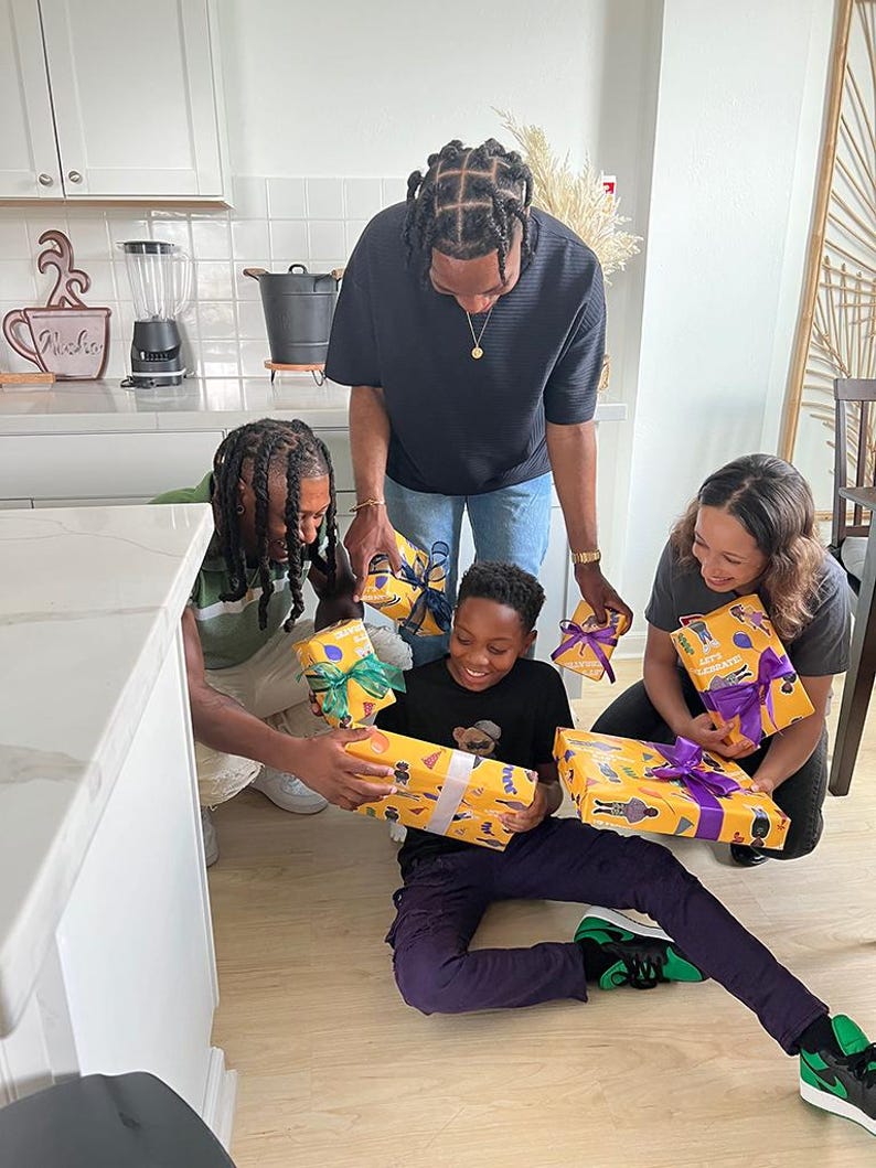 May include: A group of people are gathered, holding and admiring wrapped gifts. The gifts are wrapped in yellow paper with a repeating pattern and tied with colorful ribbons. The scene takes place in a well-lit kitchen setting.