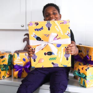 May include: A young person holds a large, yellow gift box wrapped in patterned paper with the words "LET'S CELEBRATE!" and illustrations. Several other wrapped gifts with colorful ribbons are visible.