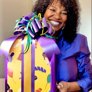 May include: A wrapped gift box with a yellow background and purple accents. The gift box is decorated with images of people, balloons, and streamers. The box is wrapped with a large purple bow and ribbon.