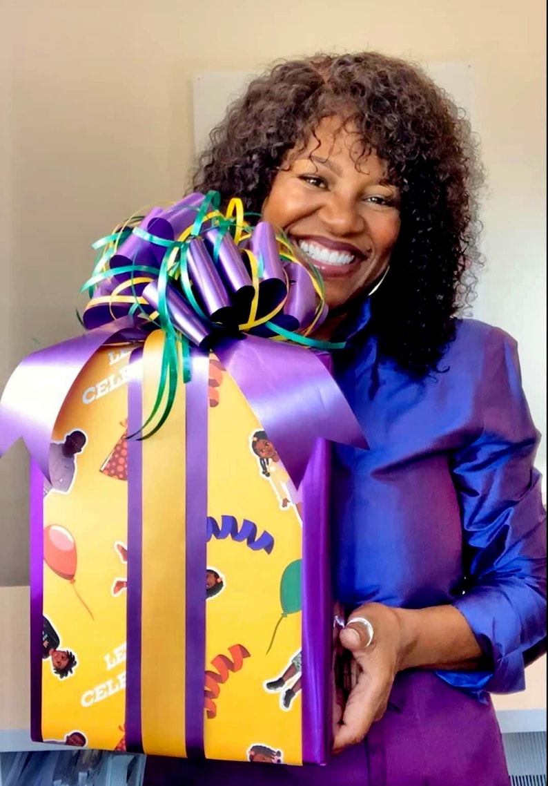 May include: A wrapped gift box with a yellow background and purple accents. The gift box is decorated with images of people, balloons, and streamers. The box is wrapped with a large purple bow and ribbon.