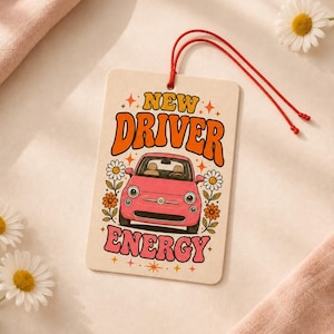 May include: A rectangular air freshener with a cream background, featuring a pink car illustration, orange and pink text reading NEW DRIVER ENERGY, and colourful daisy flowers. A thin red string is attached to the top for hanging purposes.