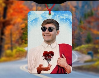 May include: A car air freshener with a photo of a person wearing sunglasses and a red robe. The person is holding a broken heart in their hand. The background is a blue sky with clouds.