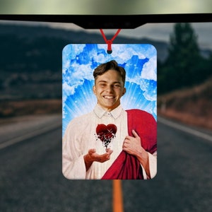 May include: Rectangular car air freshener featuring a smiling person with a broken heart design. The background includes a blue sky with clouds and sun rays. The air freshener hangs from a red string.