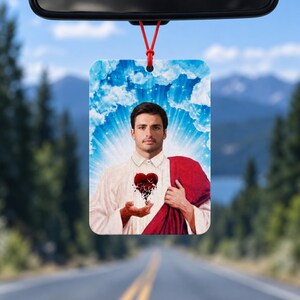 May include: A rectangular car air freshener featuring a portrait of a man with dark hair and a beard, wearing a white shirt and a red drape. He holds a stylized, dark, fragmented heart in his hands against a background of blue sky and clouds.