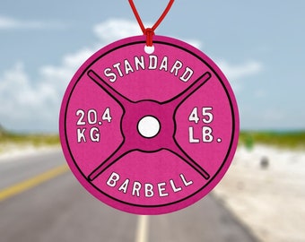Puede incluir: Un ambientador para coche con forma de pesa rosa, colgado de un espejo retrovisor. La pesa está etiquetada como "STANDARD" y muestra 20,4 kg y 45 lb. El fondo es una carretera y una playa.