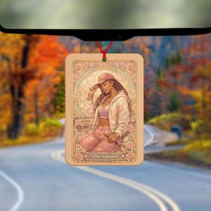 May include: Rectangular car air freshener featuring an illustration of a woman in a pink outfit and a cap, holding a microphone. The design includes roses and the text "The Queen". The air freshener hangs from a red string.
