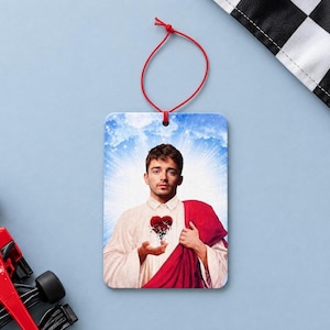May include: A rectangular hanging air freshener featuring a graphic print of a man with brown hair wearing a white shirt and a red robe, holding a cracked red heart in his hands against a blue sky background with a white sunburst effect.