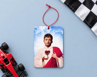 May include: A rectangular hanging air freshener featuring a graphic print of a man with brown hair wearing a white shirt and a red robe, holding a cracked red heart in his hands against a blue sky background with a white sunburst effect.