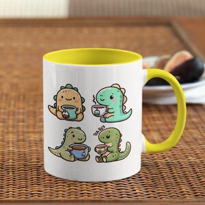 May include: A white ceramic mug with a yellow rim and handle. The mug features a cartoon illustration of four dinosaurs drinking tea. The text "Tea-Rex" is printed on the mug.