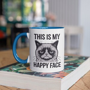 May include: White ceramic mug with a blue rim and handle. The mug features a black and white illustration of a grumpy cat with the text "This is my happy face".