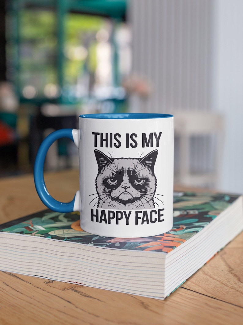 May include: White ceramic mug with a blue rim and handle. The mug features a black and white illustration of a grumpy cat with the text "This is my happy face".