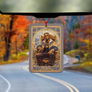 May include: Rectangular car air freshener featuring an illustration of a person wearing a crown and sitting on a throne. The text "The King" and "Hip Hop" are at the bottom. The air freshener is hanging from a red string.