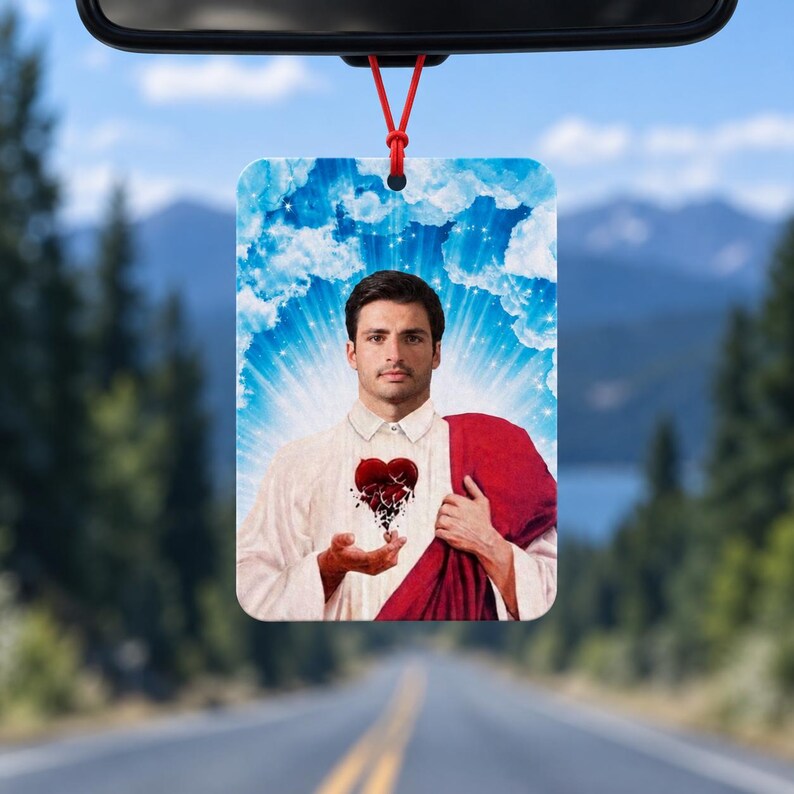 May include: A rectangular car air freshener featuring a portrait of a man with dark hair and a beard, wearing a white shirt and a red drape. He holds a stylized, dark, fragmented heart in his hands against a background of blue sky and clouds.