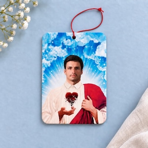 May include: A rectangular air freshener featuring a portrait of a person with dark hair and a mustache, wearing a white collared shirt and a red drape. A dark red heart graphic is centered on the chest. The background shows blue sky and clouds.