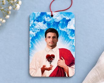 May include: A rectangular air freshener featuring a portrait of a person with dark hair and a mustache, wearing a white collared shirt and a red drape. A dark red heart graphic is centered on the chest. The background shows blue sky and clouds.