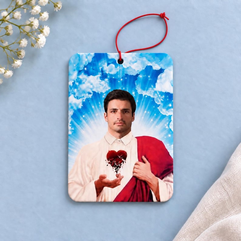 May include: A rectangular air freshener featuring a portrait of a person with dark hair and a mustache, wearing a white collared shirt and a red drape. A dark red heart graphic is centered on the chest. The background shows blue sky and clouds.