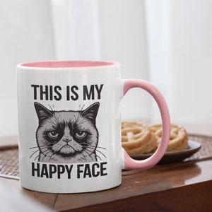 May include: White ceramic mug with a pink rim and handle. The mug has a black and white illustration of a grumpy cat with the text "This is my happy face" printed on it.