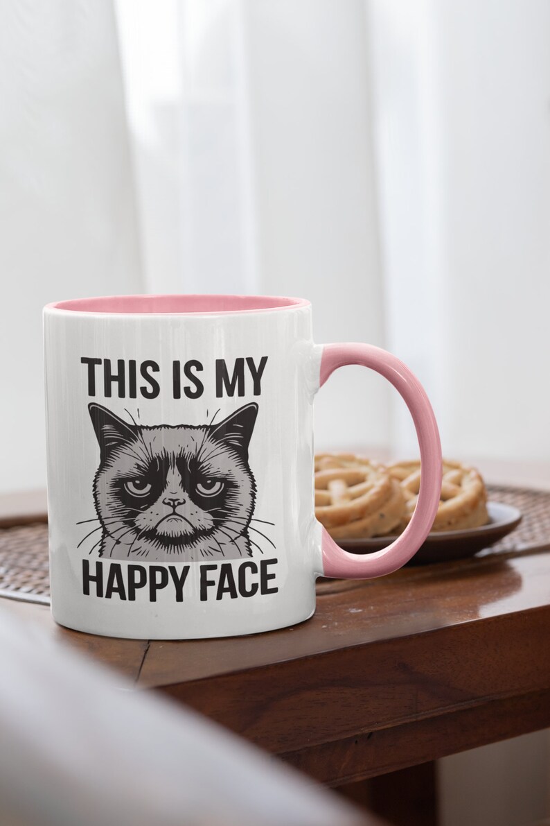 May include: White ceramic mug with a pink rim and handle. The mug has a black and white illustration of a grumpy cat with the text "This is my happy face" printed on it.