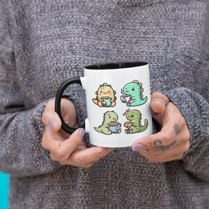 May include: White ceramic mug with a black rim and handle. The mug features a cartoon illustration of four green dinosaurs drinking tea. The text "Tea Rex" is printed on the mug.