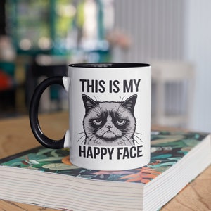 May include: White ceramic mug with a black handle and black rim. The mug features a black and white illustration of a grumpy cat with the text "This is my happy face".