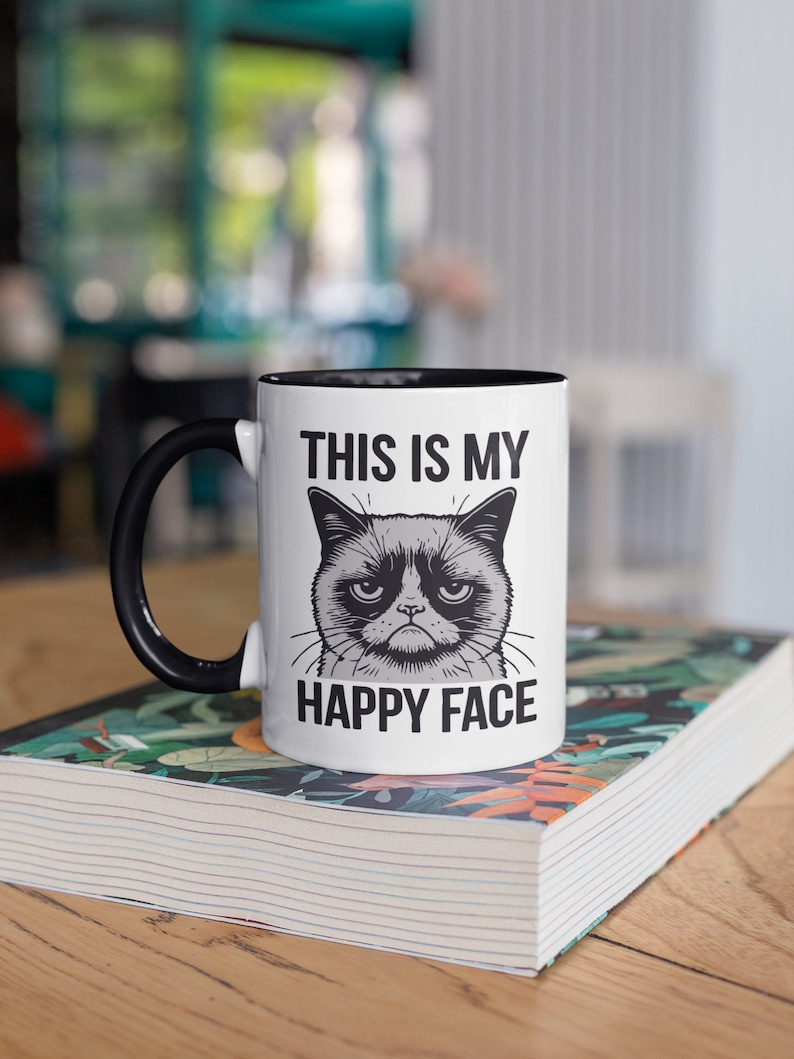May include: White ceramic mug with a black handle and black rim. The mug features a black and white illustration of a grumpy cat with the text "This is my happy face".