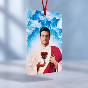 May include: A rectangular hanging ornament featuring a portrait of a man with dark hair wearing a white collared shirt and a red drape. He holds a stylized red heart that appears to be shattering into small pieces against a blue sky background.