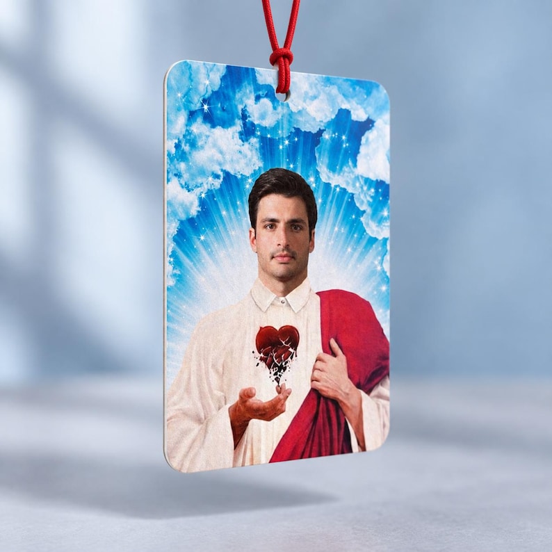 May include: A rectangular hanging ornament featuring a portrait of a man with dark hair wearing a white collared shirt and a red drape. He holds a stylized red heart that appears to be shattering into small pieces against a blue sky background.