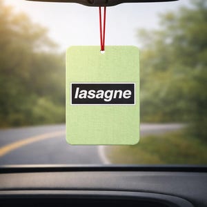 May include: A pale green rectangular car air freshener with the word "lasagne" in a black rectangle. It hangs from a red string. The background shows a road and trees.