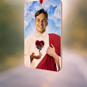 May include: Rectangular ornament with a smiling person and a broken heart design. The person wears a white shirt and red cloth. The background shows a blue sky with clouds. A red ribbon is attached for hanging.